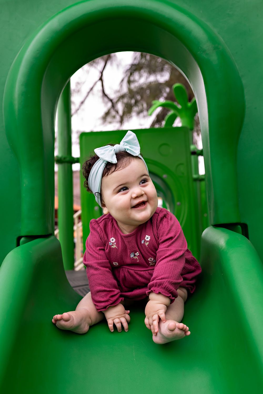 A small child wearing a burgundy outfit and bow plays happily inside a green playground slide.