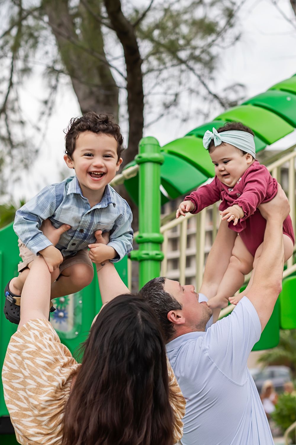 Family enjoying playful moment at playground with parents lifting children up against green play structure background.