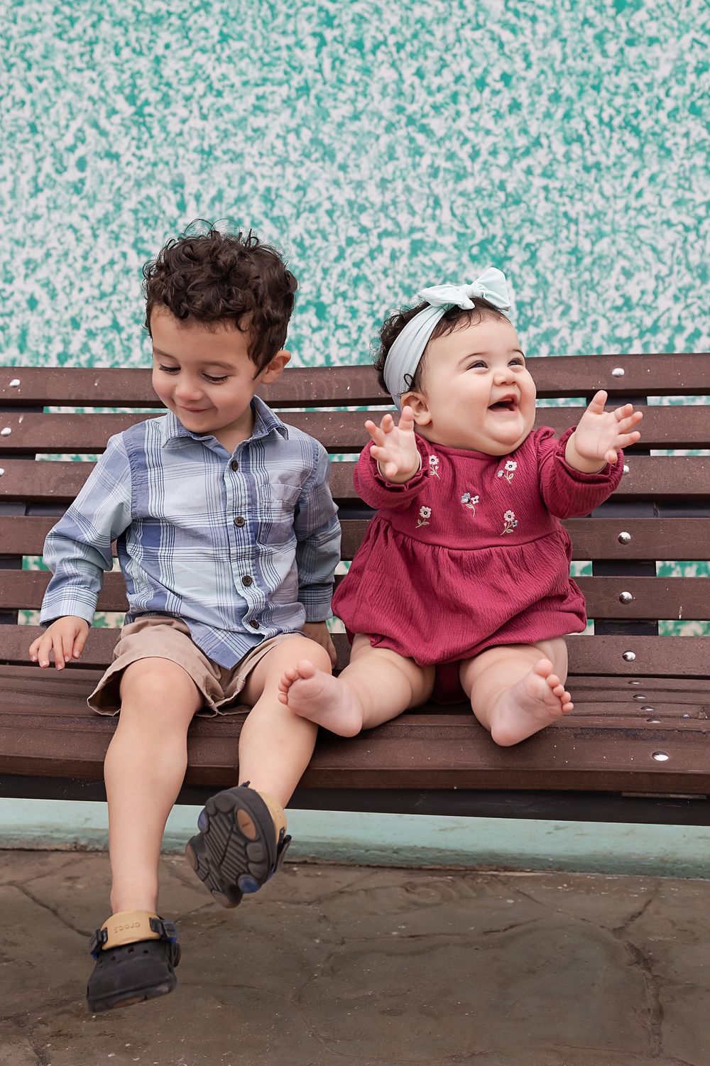 Two young children sit together on a wooden bench, laughing and playing while wearing casual clothes.