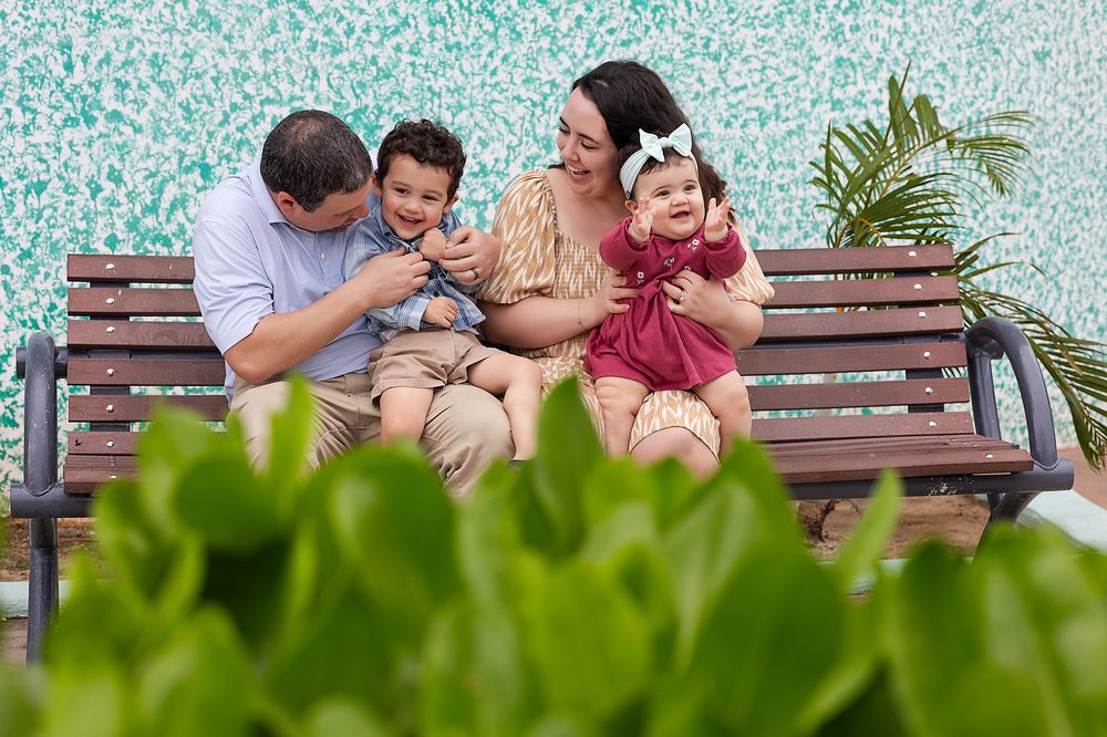 A family sits together on wooden benches in an outdoor garden setting while playing with their husky dog.