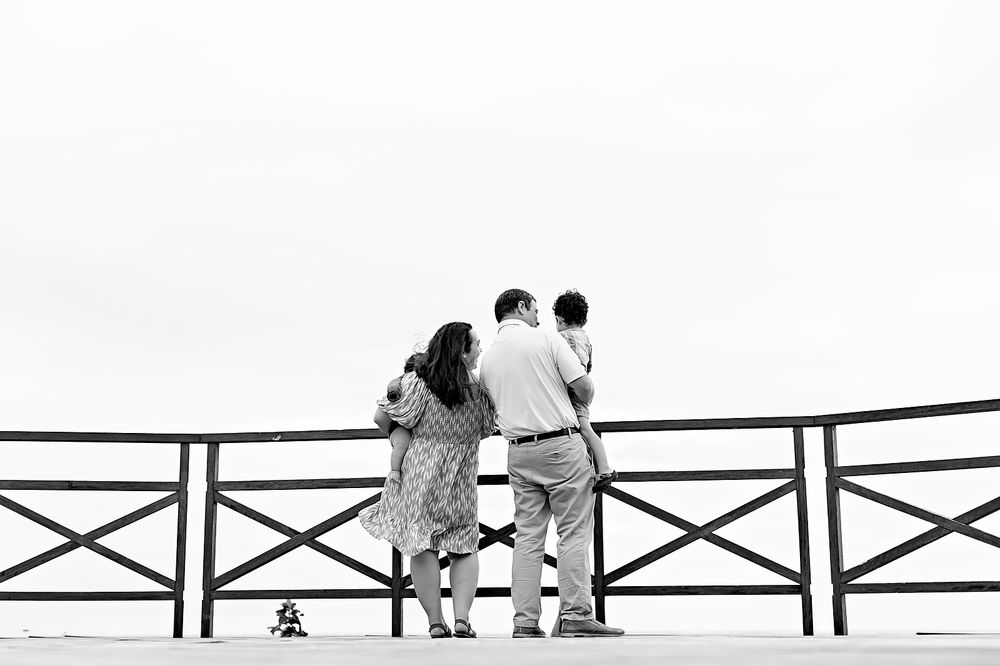 Three people sit on a wooden pier railing silhouetted against a bright white background.