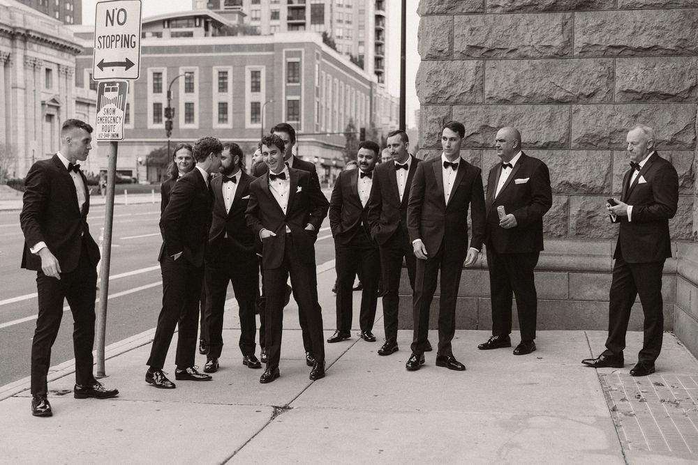 A sequence of groomsmen walking and laughing together in black tuxedos along a stone building exterior.