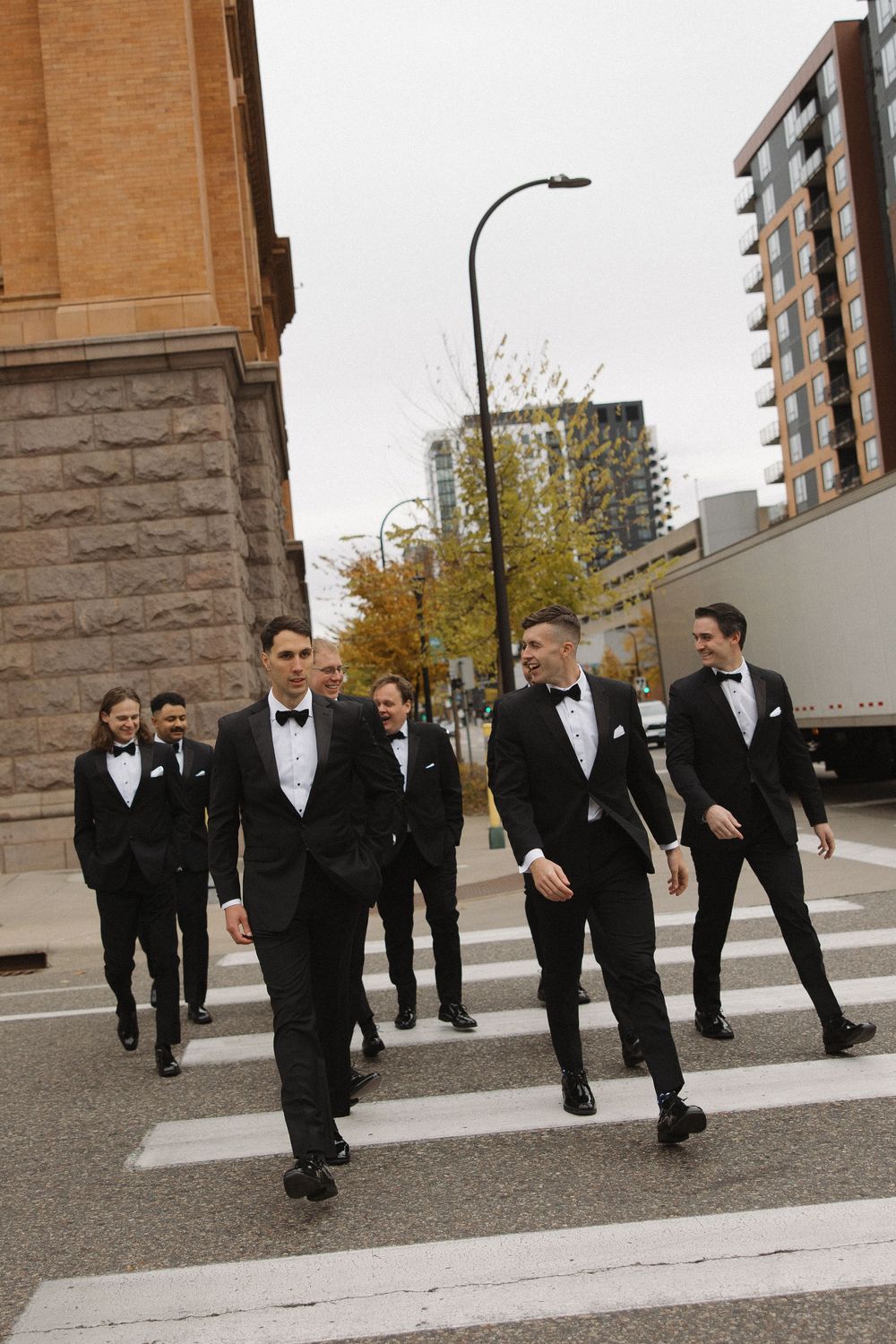 Wedding party members in formal black tuxedos walking and chatting on an urban sidewalk.