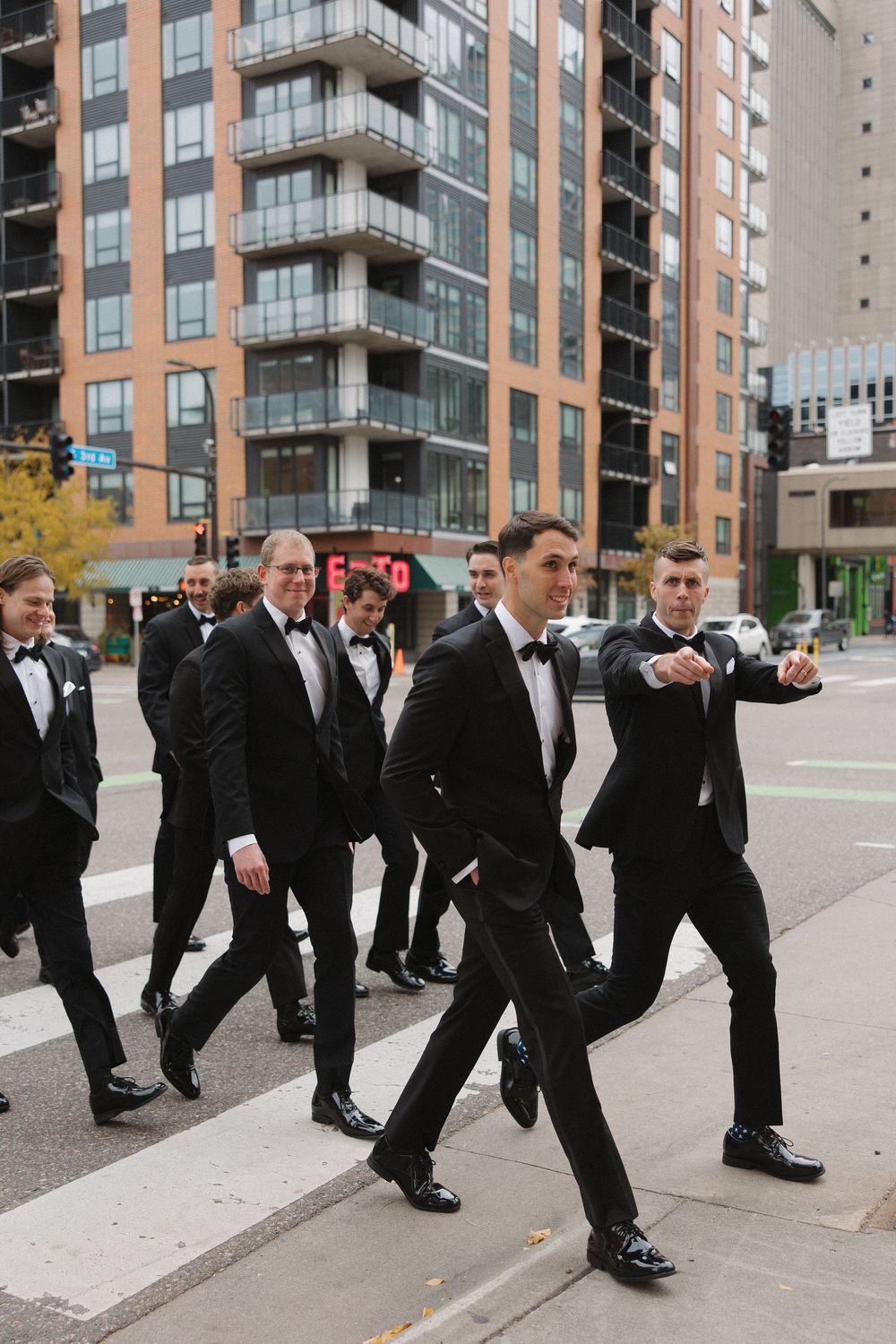 Groomsmen in black tuxedos walking in a synchronized line formation outside a modern apartment building.