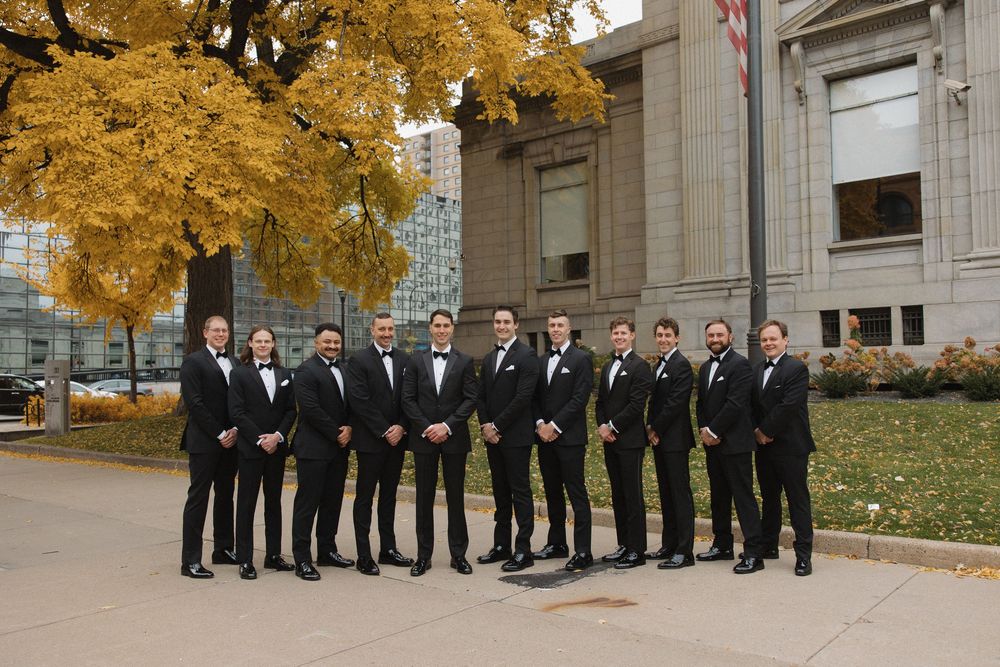 Wedding party in formal black attire posing in front of a building with golden autumn trees.