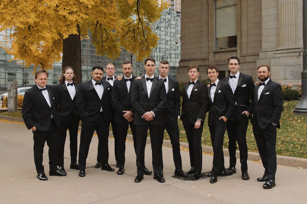 A wedding party in black tuxedos poses together on stone steps during autumn with golden leaves in the background.