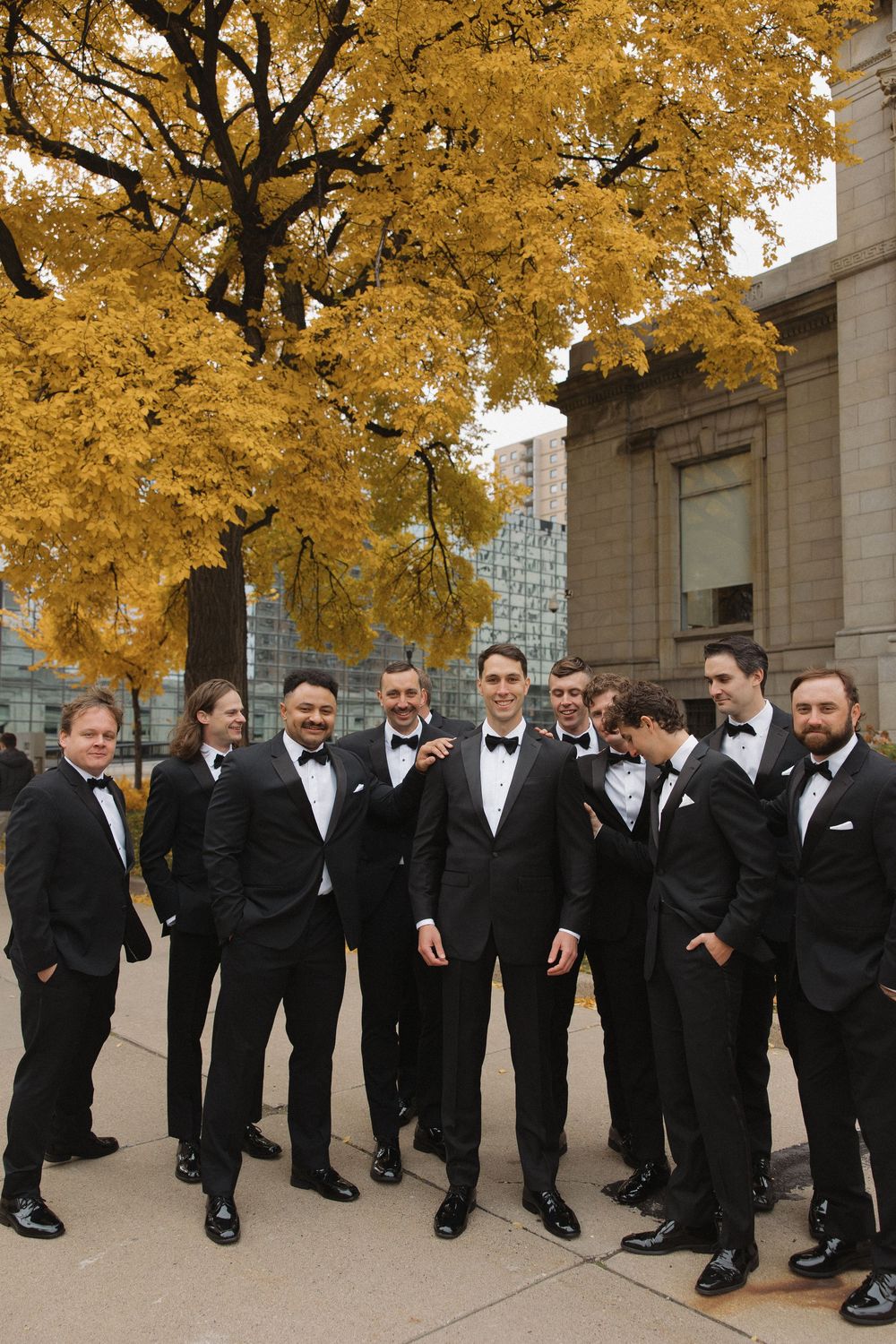 Groomsmen in black tuxedos lean against a wall under golden autumn trees during a formal wedding photo session.