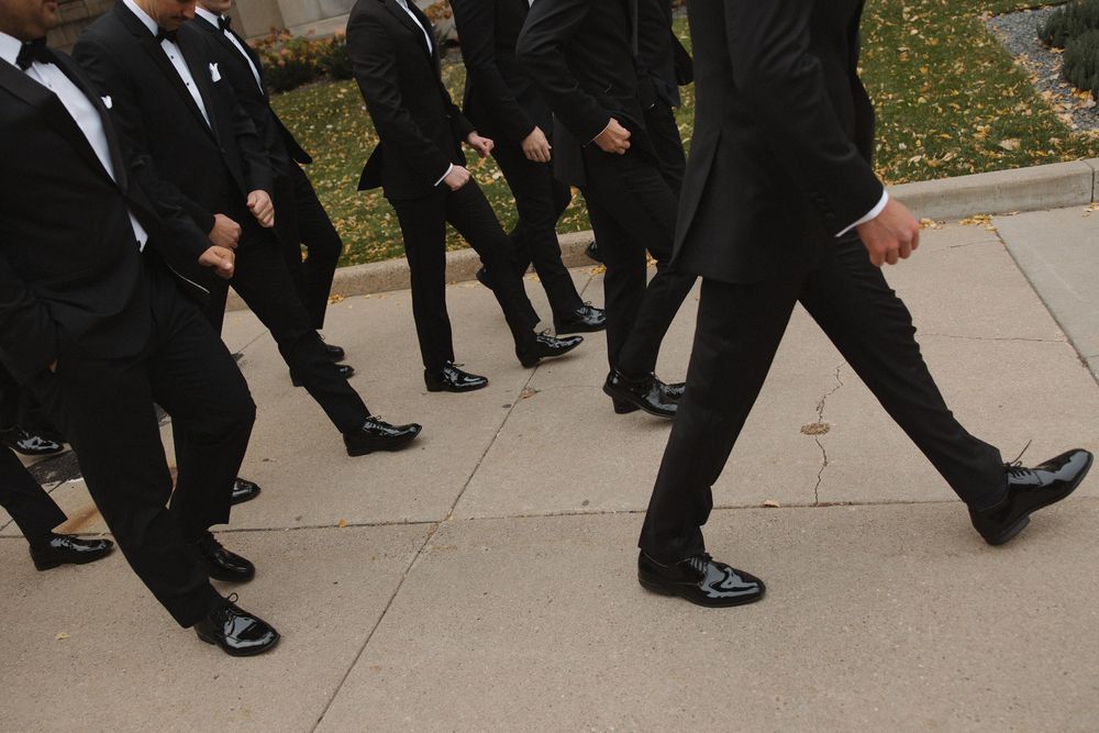 A group of people wearing formal black suits and dress shoes walking in a line on a concrete sidewalk.