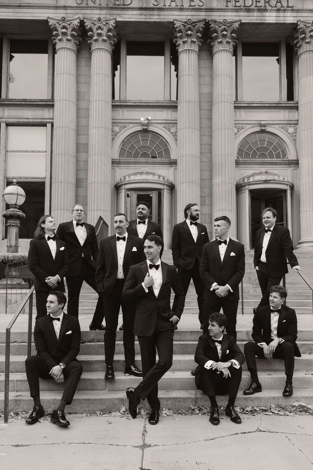 Black and white photo showing groomsmen in tuxedos posing playfully on steps of a Federal building.