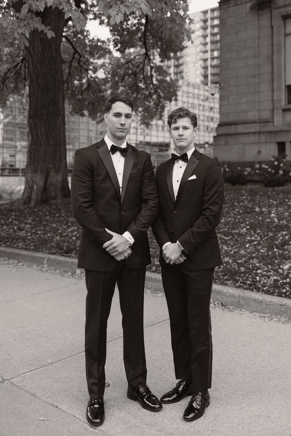 Two people in formal black suits and dress shoes stand together on a sidewalk in front of a building.
