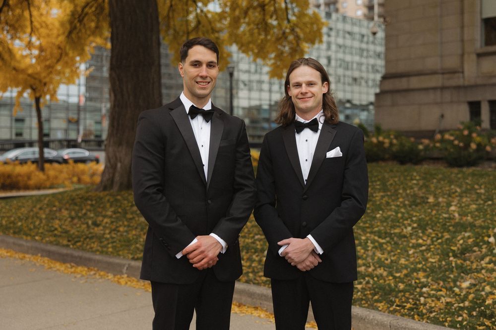 Two people in black suits stand together outdoors during autumn with golden leaves in the background.