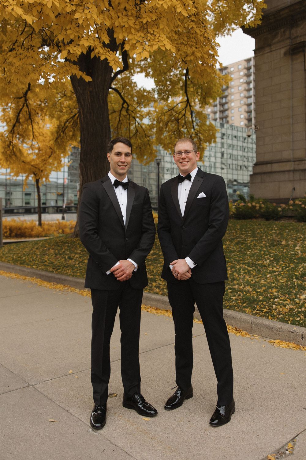 Groomsmen pose in formal black tuxedos against autumn trees with golden leaves in an urban setting.