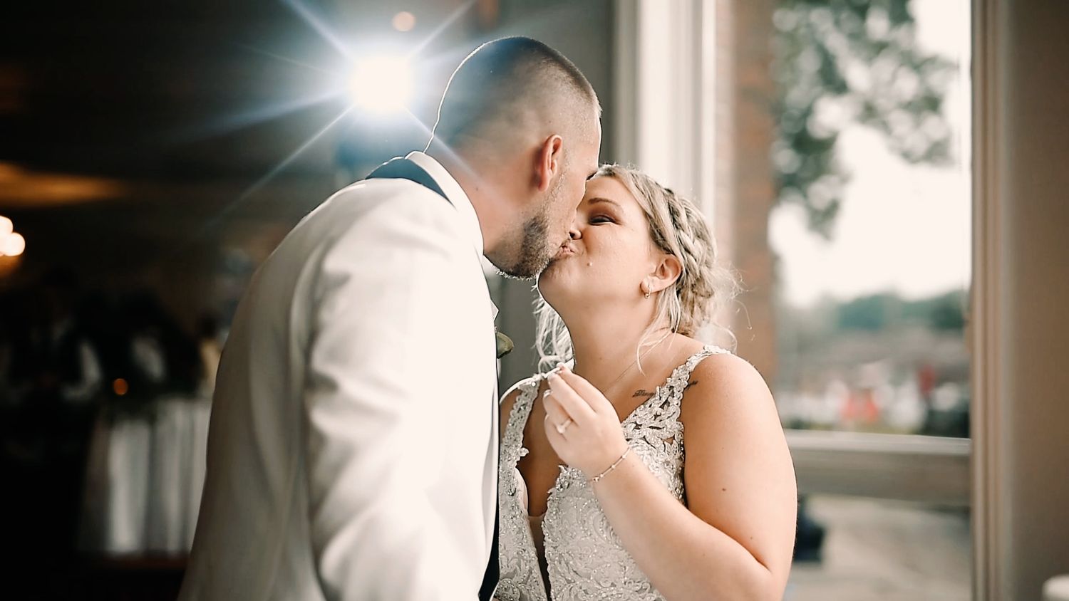 A romantic couple shares a tender kiss during their wedding reception with soft window light illuminating the scene.