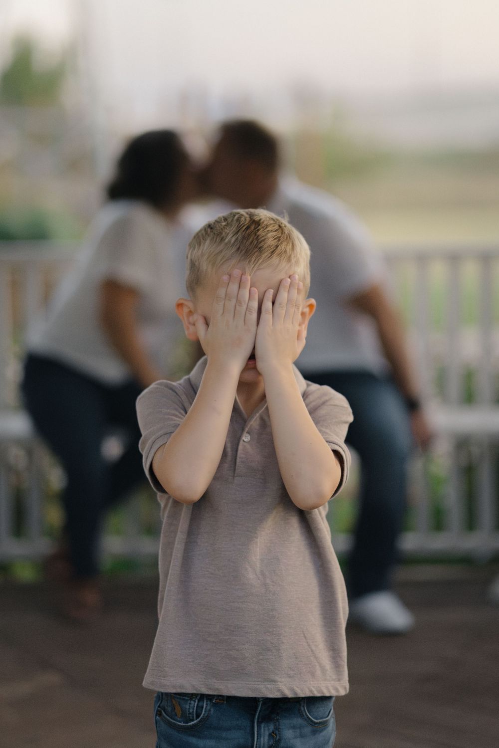 Child covers face while couple shares private moment in background on porch.