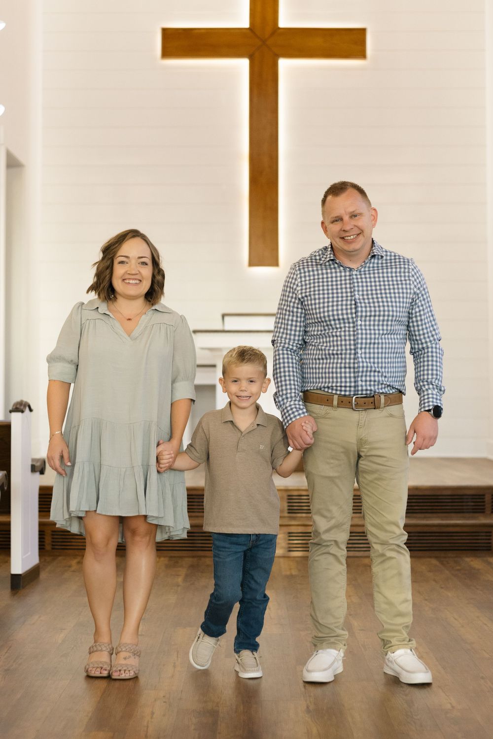 Family stands together holding hands in front of wooden cross inside church sanctuary.