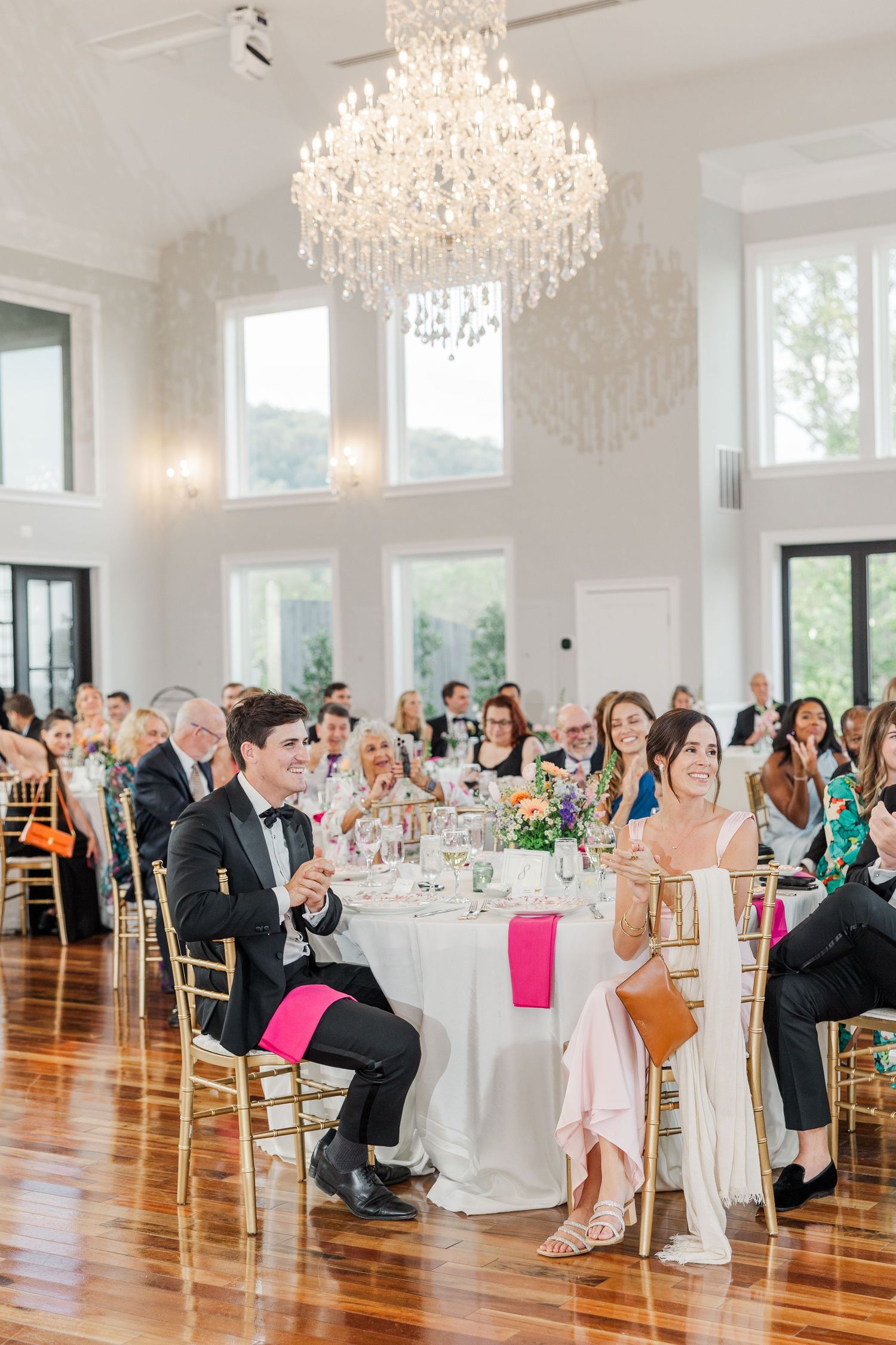 Wedding reception venue with crystal chandelier and guests seated at round tables with pink accents.