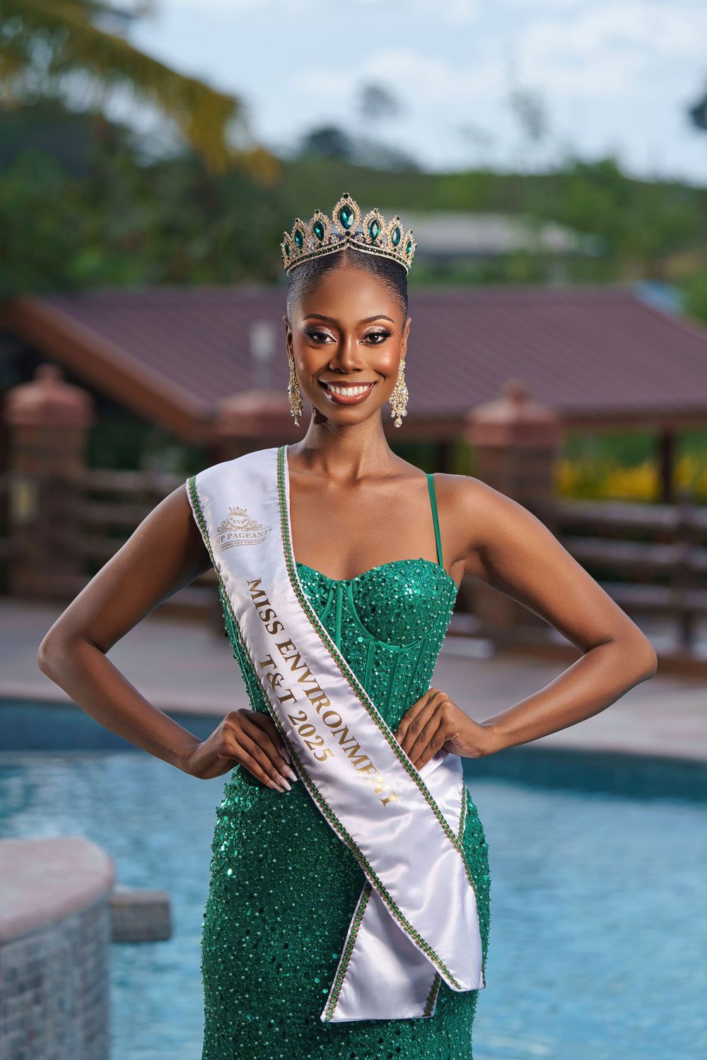 Beauty pageant winner poses in emerald green sequined gown and white sash by swimming pool.