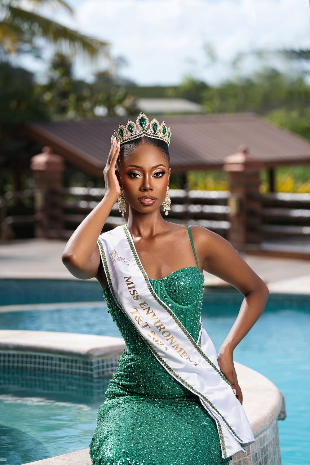 Beauty pageant winner in a sparkling emerald gown and crown poses elegantly by a swimming pool.