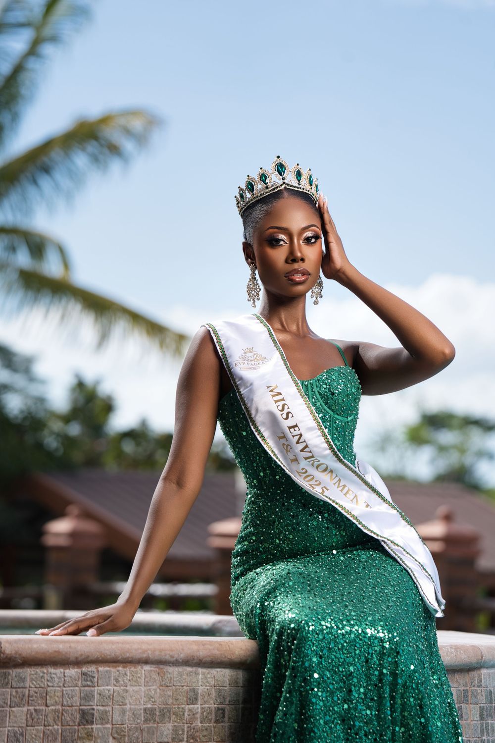 Beauty pageant winner in sparkling green gown and crown poses elegantly against tropical backdrop with palm trees.