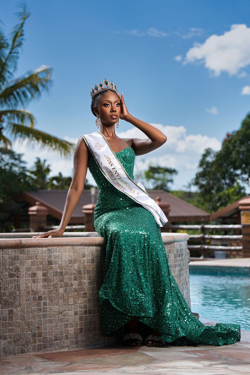 Pageant winner in a glittering green evening dress poses gracefully beside a resort pool.