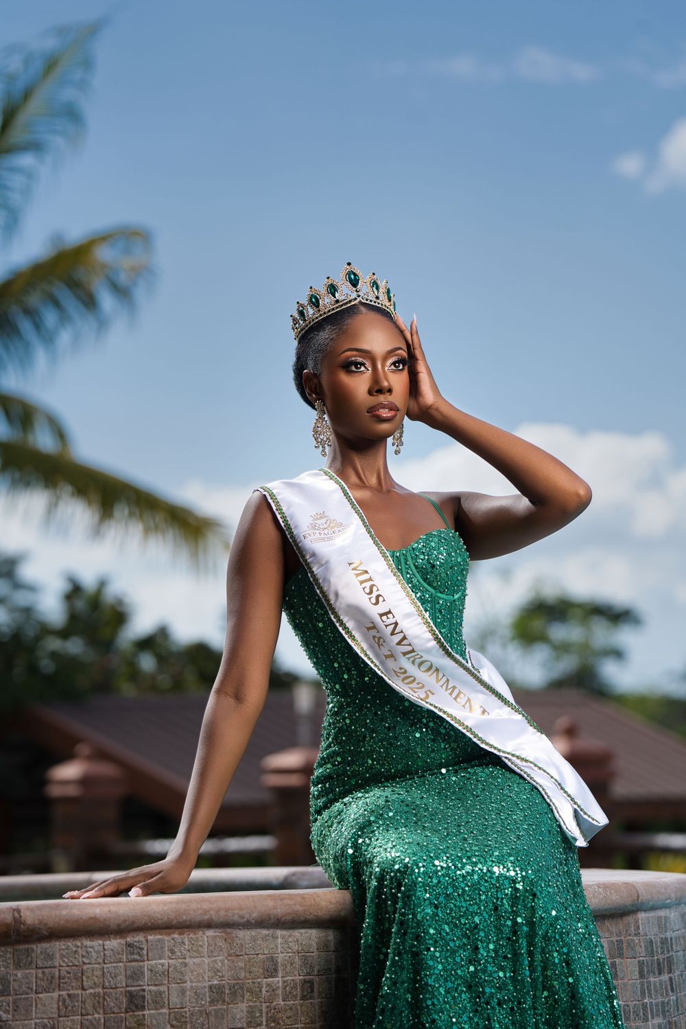 A woman in a sparkling emerald green gown and pageant sash poses gracefully against a stone wall with palm trees behind.