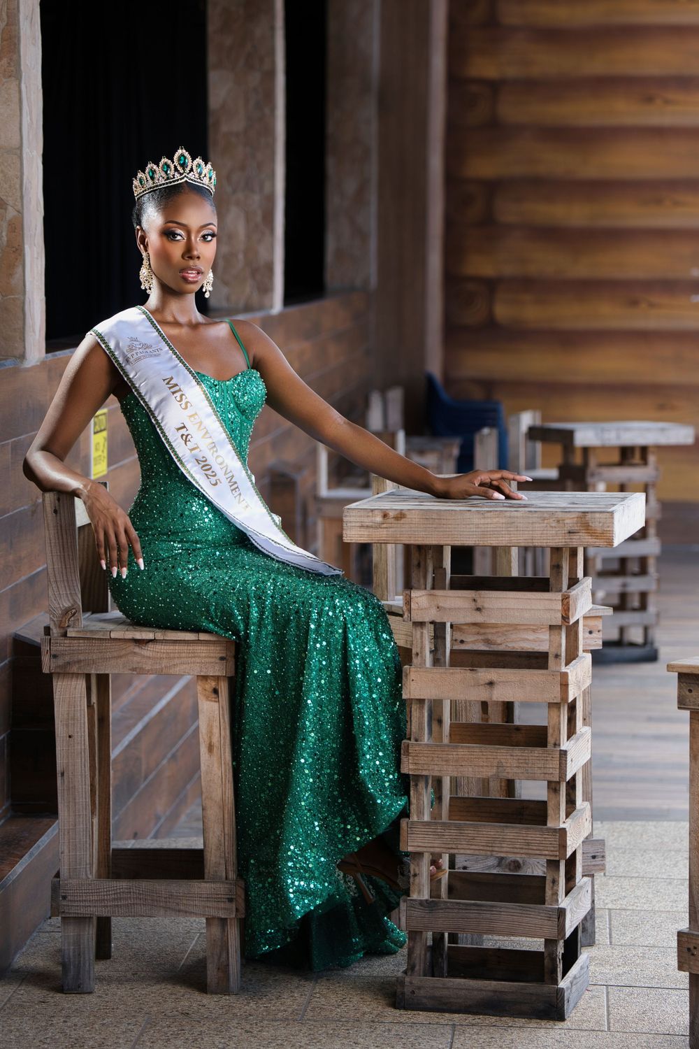 Beauty pageant contestant in sparkling green gown poses elegantly on wooden chairs in rustic setting.