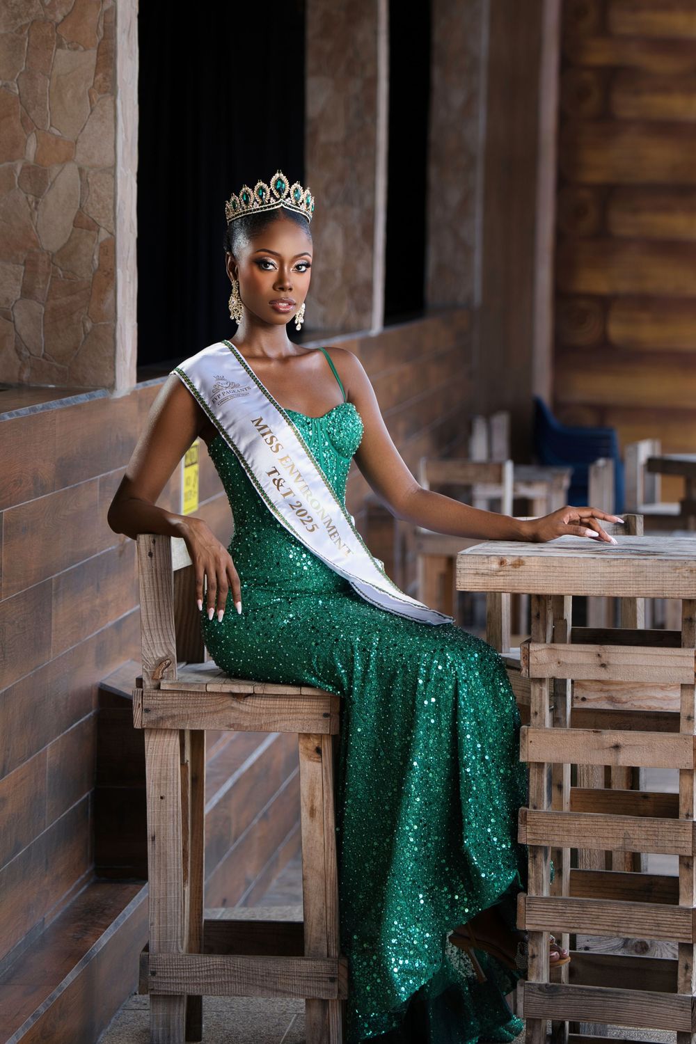 A pageant winner in a sparkling emerald gown and crown poses gracefully on wooden benches in a rustic setting.