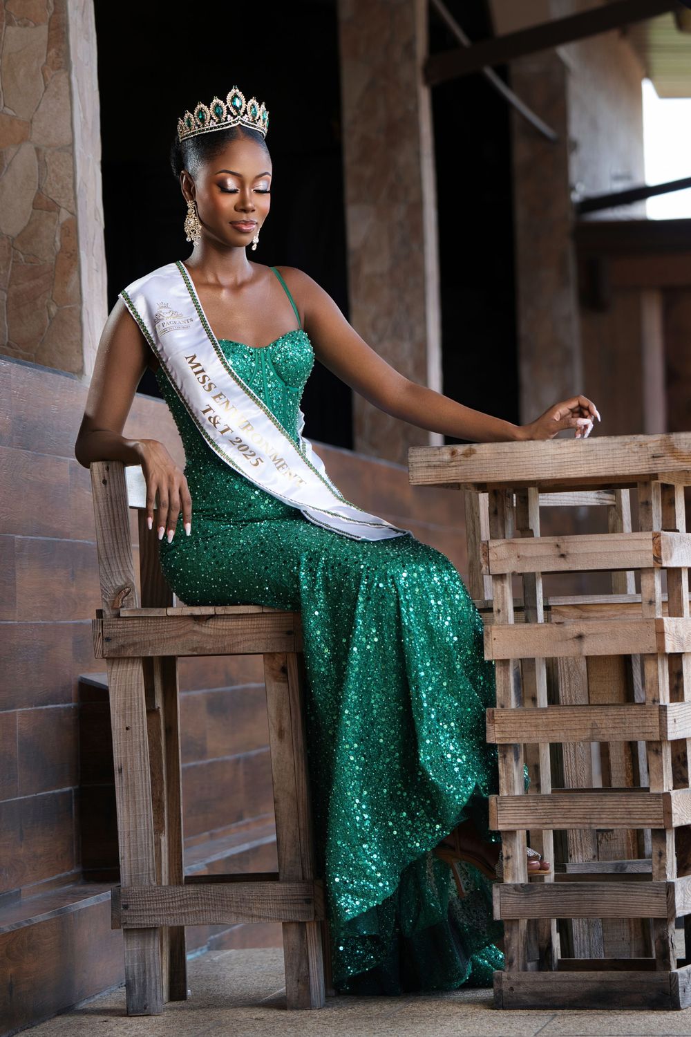 A beauty queen in a glittering green dress sits elegantly on wooden seating against a stone wall backdrop.