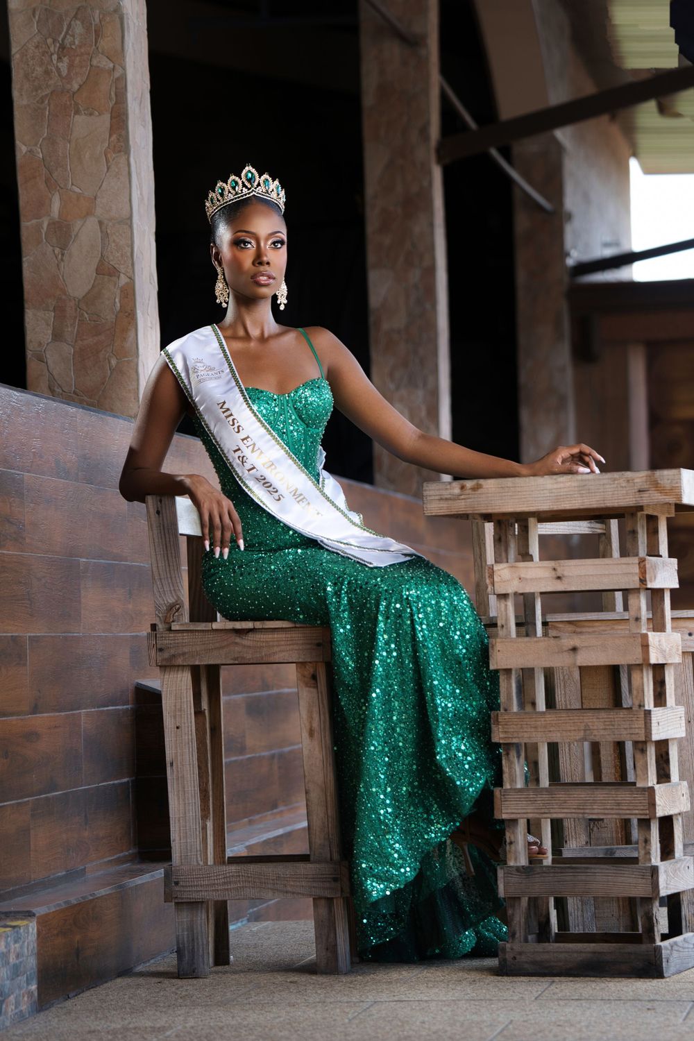 A pageant winner in a sparkling emerald green gown and tiara sits elegantly on wooden stairs.