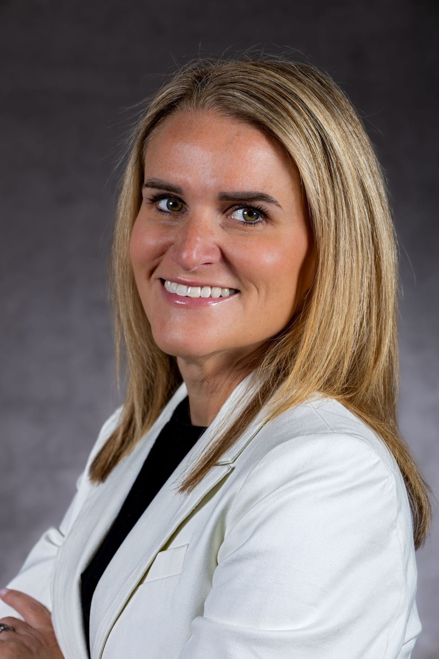 Professional headshot portrait of person in white blazer with black lapel against dark gray background.