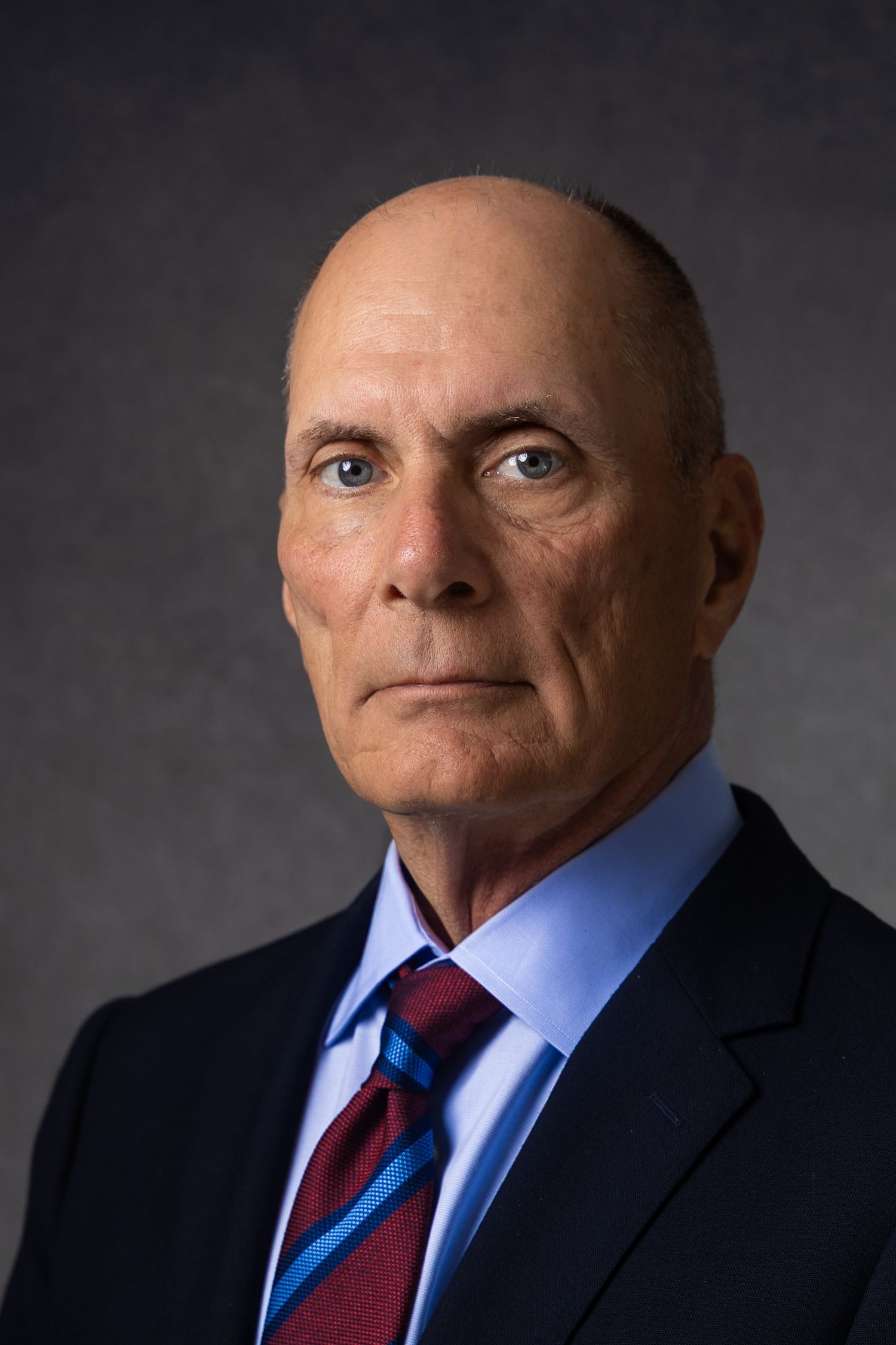 Professional headshot of a bald middle-aged man in a navy suit and striped tie against gray background.