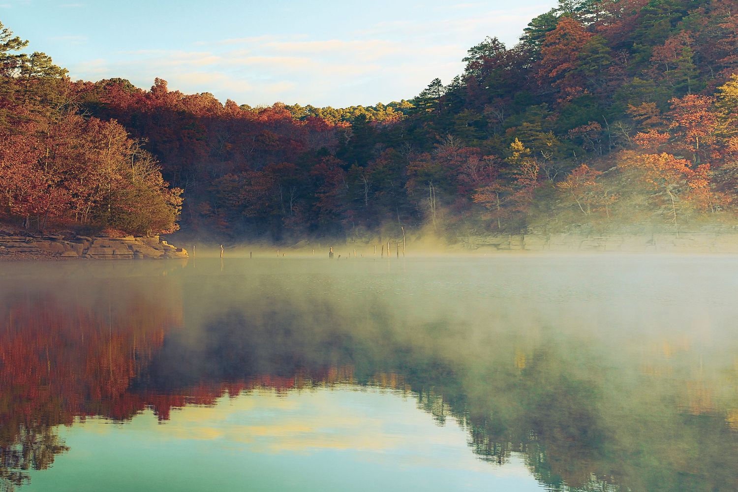 Misty lake surrounded by autumn trees with orange and red foliage reflecting in calm waters at sunrise.