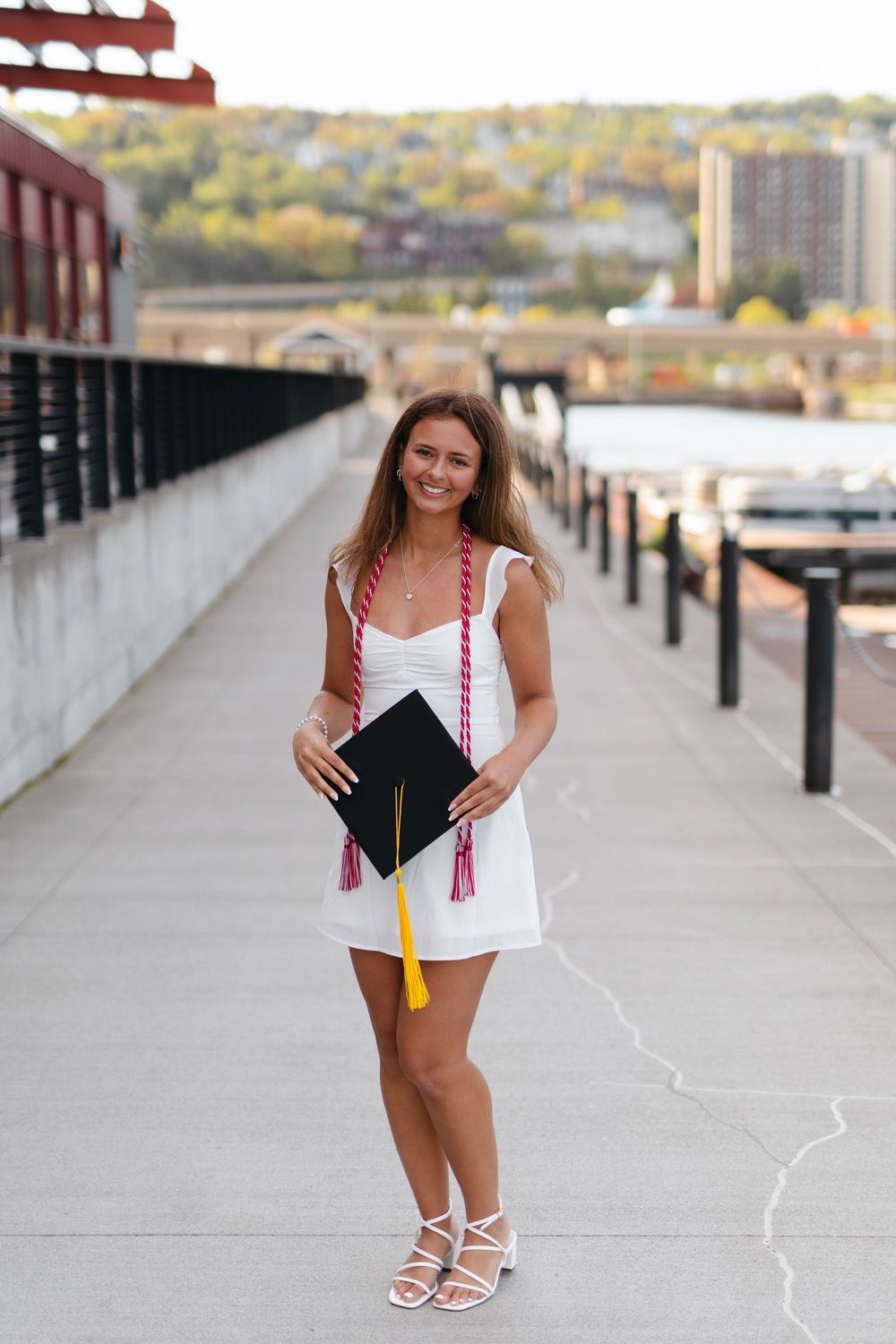 Graduate poses with decorated mortarboard on concrete pathway with urban skyline in background.