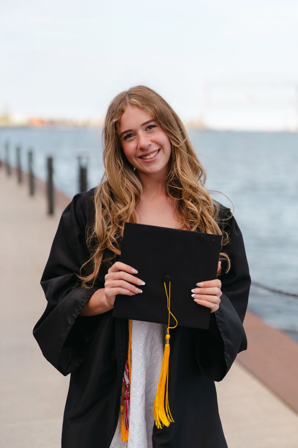 A graduate in a black cap and gown smiles while posing for photos by a waterfront pier during sunset.