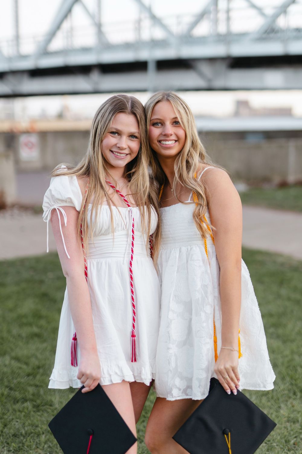 Two graduates in white dresses display their decorated mortarboards against bridge backdrop.