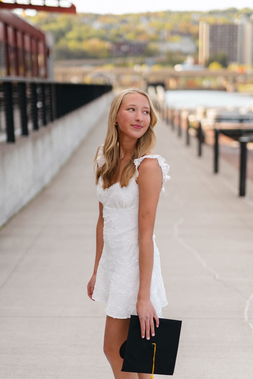 A graduate in a white lace dress holds a diploma while standing on an outdoor walkway at sunset.