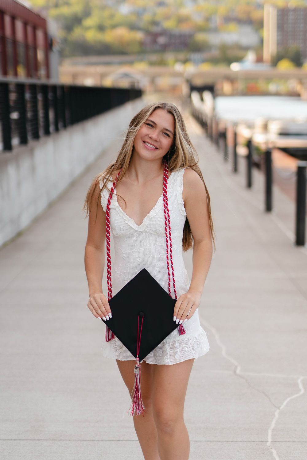 Graduate in white dress holding decorated graduation cap on urban walkway with city backdrop.