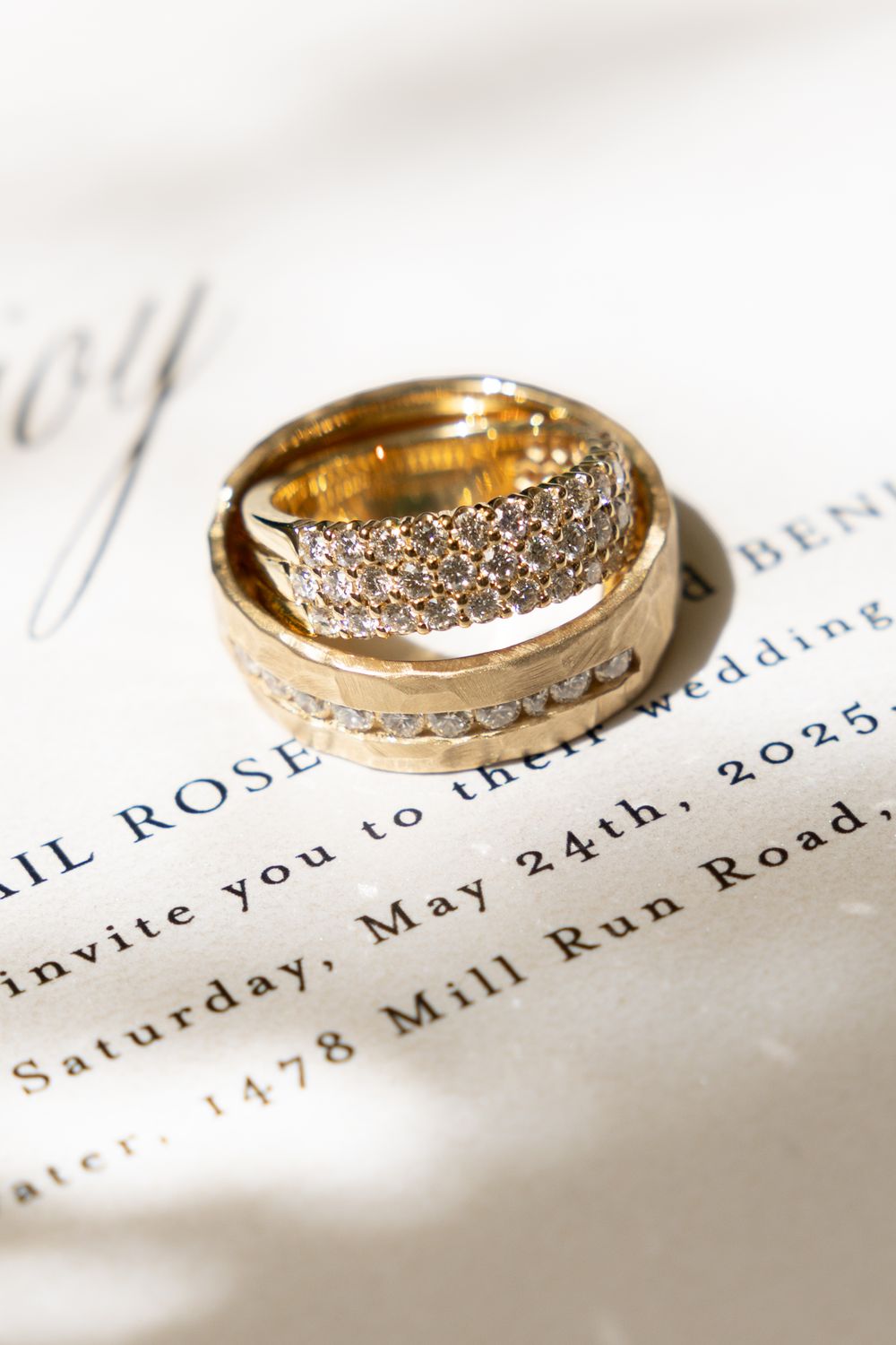 Two gold wedding rings with diamond accents resting on an elegant wedding invitation with text details.