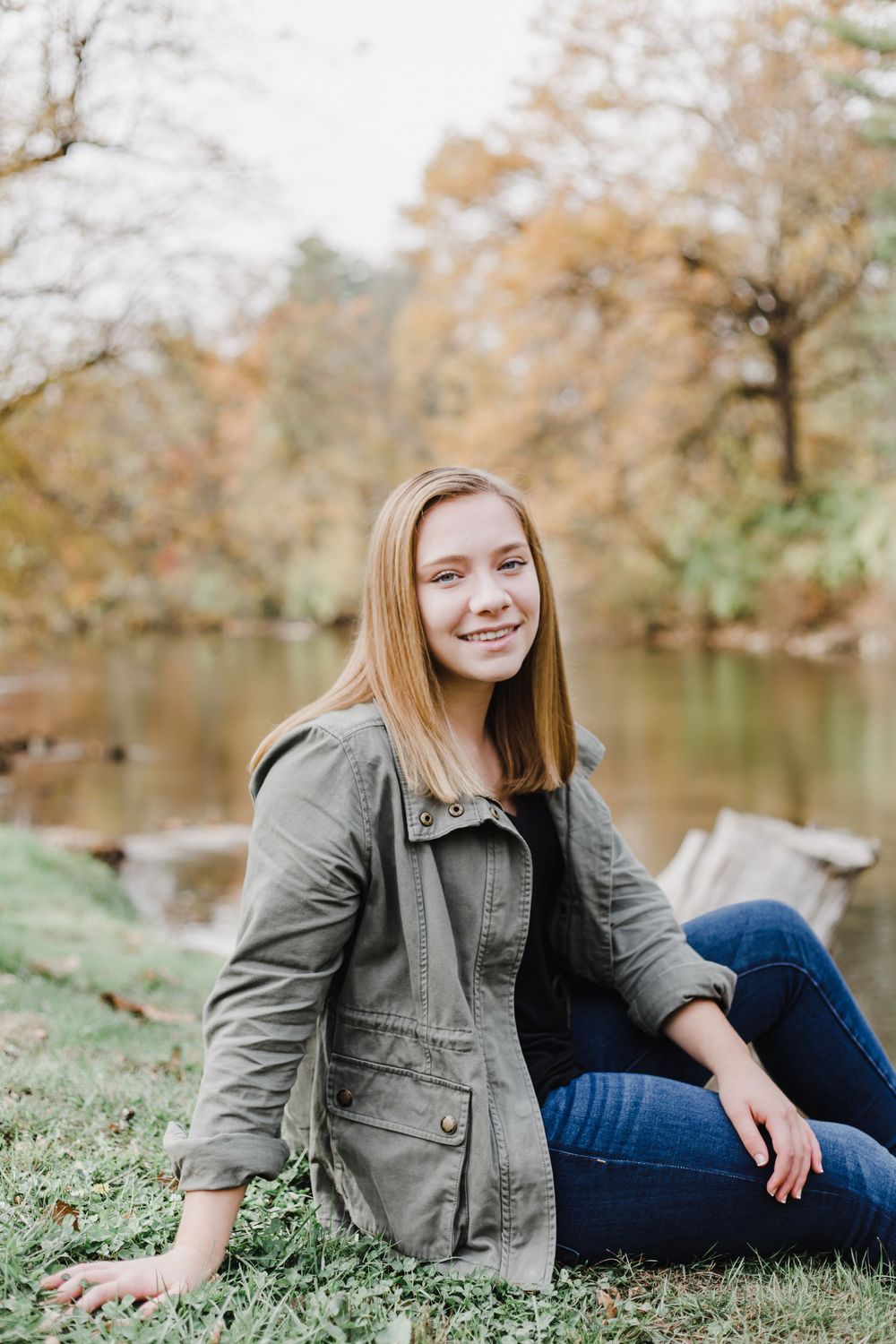 A casual outdoor portrait by a lake in autumn with fallen leaves and trees in the background.
