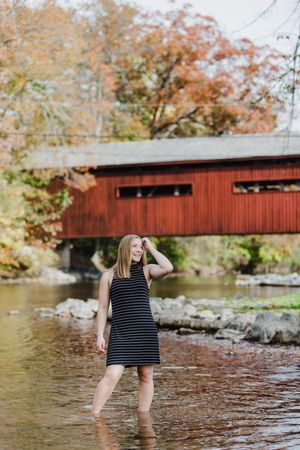 Person wading in stream water near red covered bridge during fall season.