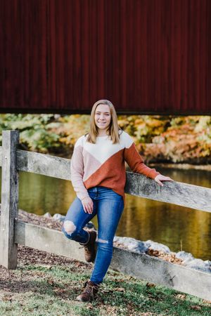 Casual autumn portrait by red barn with wooden fence and fall foliage in background.