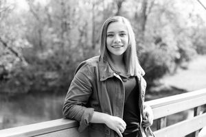 Black and white portrait of someone in a jacket standing on a bridge over water with trees in the background.