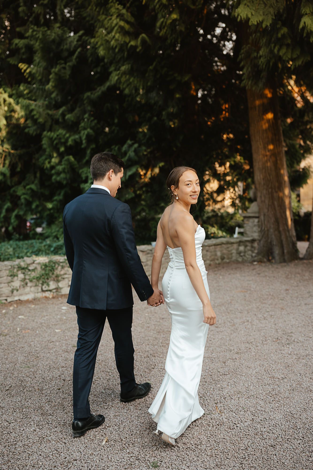 A couple in wedding attire walking hand in hand on a gravel path surrounded by trees at sunset.