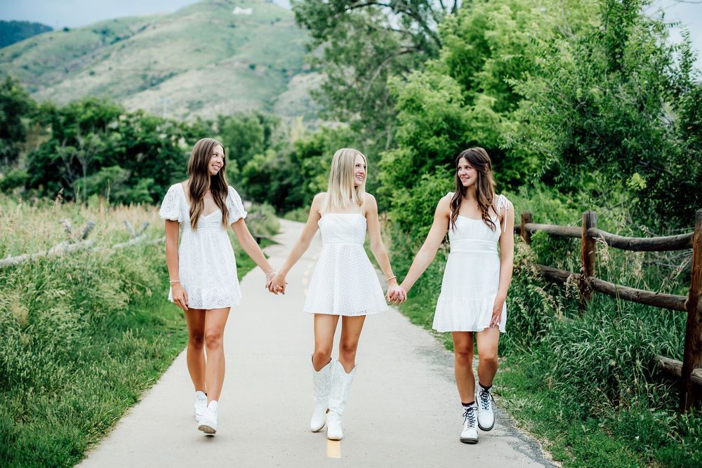 Three people in white dresses walk together on a grassy mountain path holding hands.