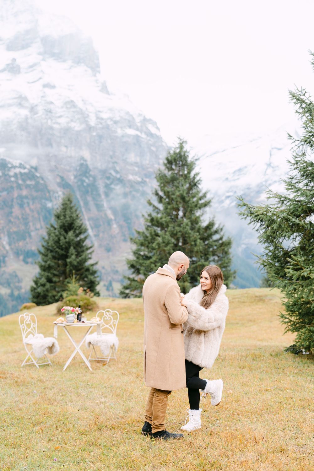 A romantic mountain proposal scene unfolds on a grassy hillside with white chairs and evergreen trees in the background.