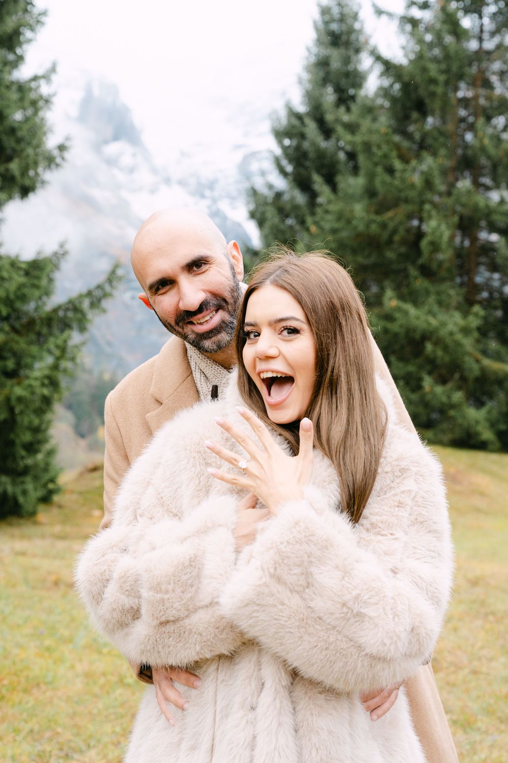 A couple wearing matching cream sweaters share playful moments together in a mountain meadow with evergreen trees behind them.