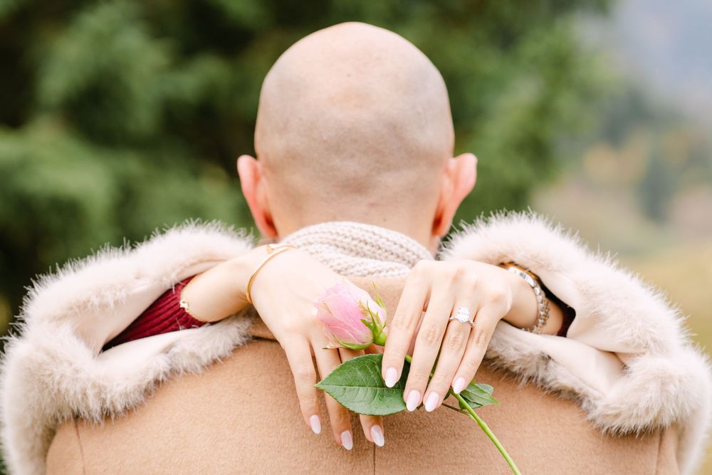 A person wearing a pink fur coat and green nail polish holds up a delicate leaf against a blurred outdoor background.