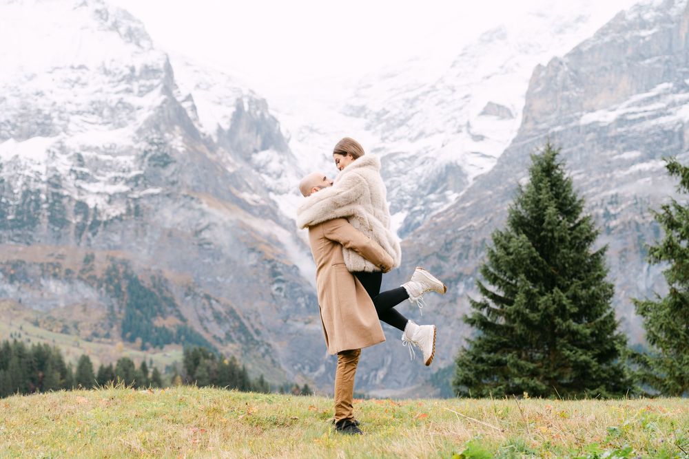 Person in a tan coat spins joyfully in an alpine meadow with snow-capped mountain peaks in the background.