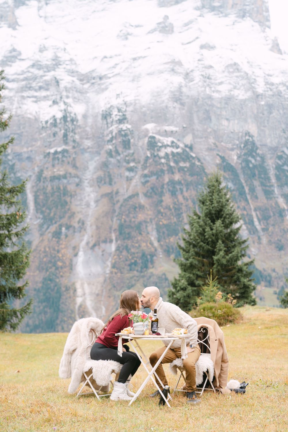 Two people share an intimate moment at a rustic table in the Alps, surrounded by majestic snow-capped mountains.
