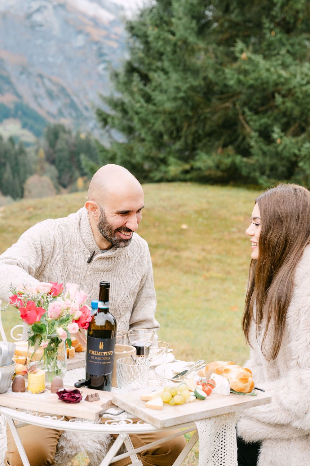 A romantic outdoor picnic setup in the mountains with a white table, drinks, and floral arrangements.